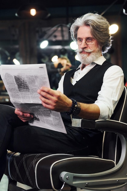 Lettura del giornale l'uomo anziano moderno ed elegante con i capelli grigi e la barba è al chiuso