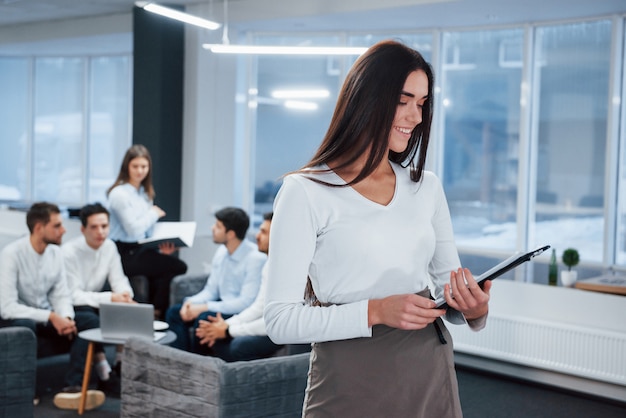 Reading the report. Portrait of young girl stands in the office with employees at background