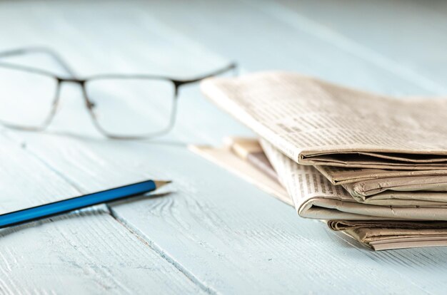 写真 朝のニュースを読む朝日に照らされた木のテーブルの上に山積みの新聞グラスと鉛筆