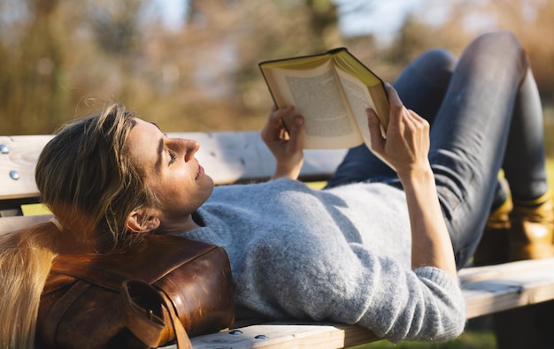 本のコンセプト画像を読んでいるベンチの学生で彼女のバッグに公園で横たわっている読書の女性