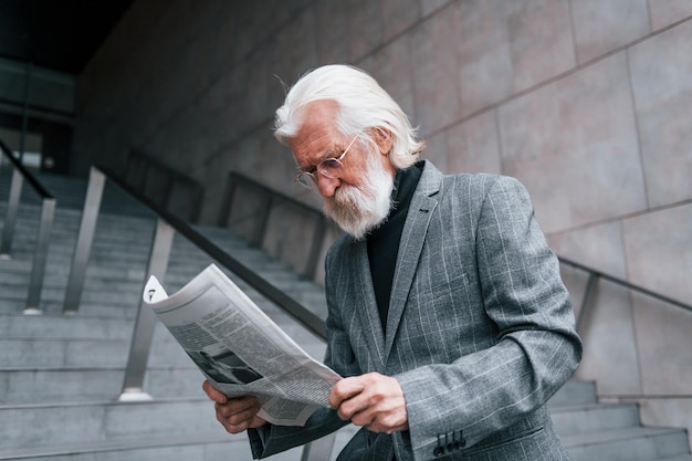 Reads newspaper Senior businessman in formal clothes with grey hair and beard is outdoors