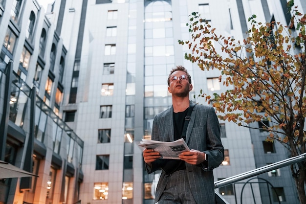 Reads newspaper Young elegant man in good clothes is outdoors in the city at daytime