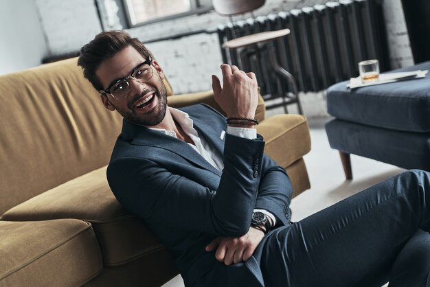 Ready to break hearts. Handsome young man in full suit smiling and looking away while sitting on the floor at home