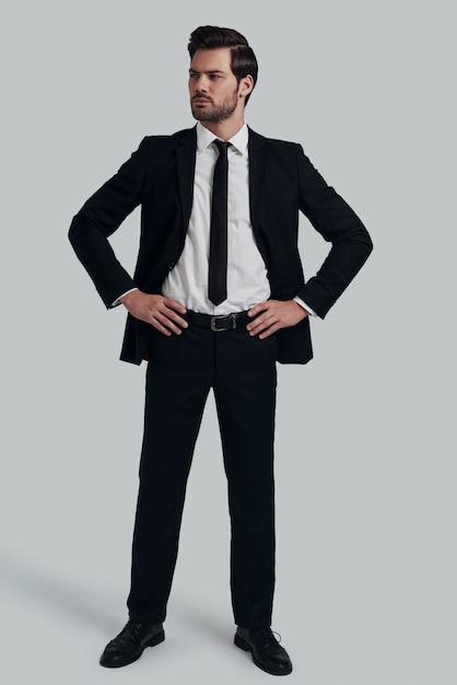 Ready to do business. Full length of handsome young man in full suit looking at camera and keeping hands on hips while standing against grey background