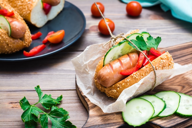 Ready-to-eat appetizing hot dog made from fried sausage, rolls and fresh vegetables, wrapped in parchment paper on a cutting board on a wooden table