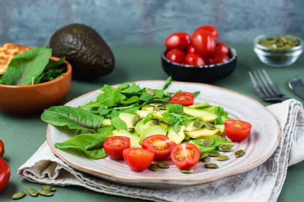 Ready-to-eat salad with avocado, tomatoes, arugula leaves, mizuna, chard and pumpkin seeds on a plate on a green background. Vegetarian vitamin food.