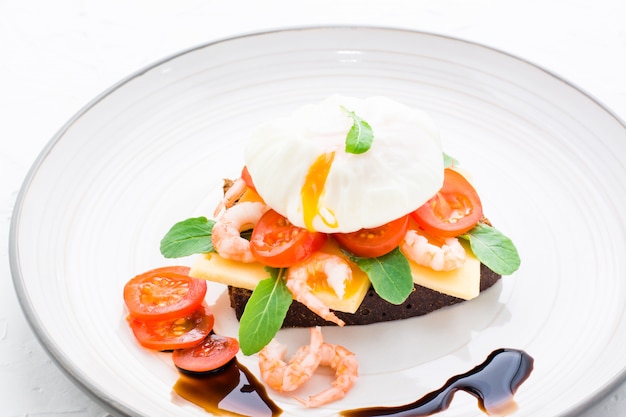 Ready-to-eat sandwich on rye bread with poached egg, tomato, shrimp and arugula on a plate