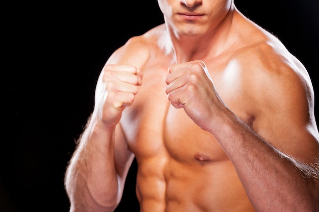 Ready to fight. Close-up of serious young shirtless man looking at camera while standing against black background