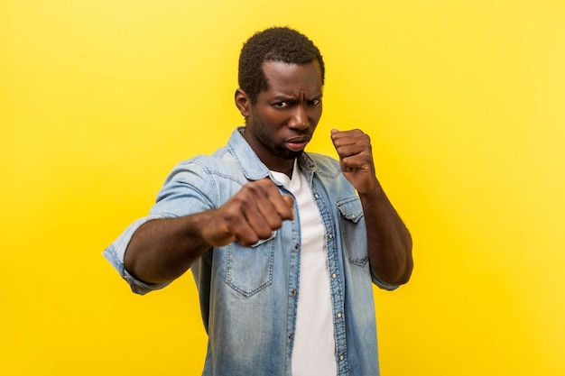 Ready for fight. Portrait of aggressive man in denim shirt with rolled up sleeves standing in defensive posture with clenched fists, threatening to attack. indoor studio shot isolated on yellow
