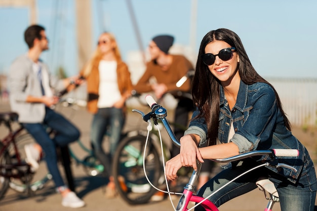 Ready to fun and adventures. Beautiful young smiling woman leaning at her bicycle and looking at camera while her friends talking in the background