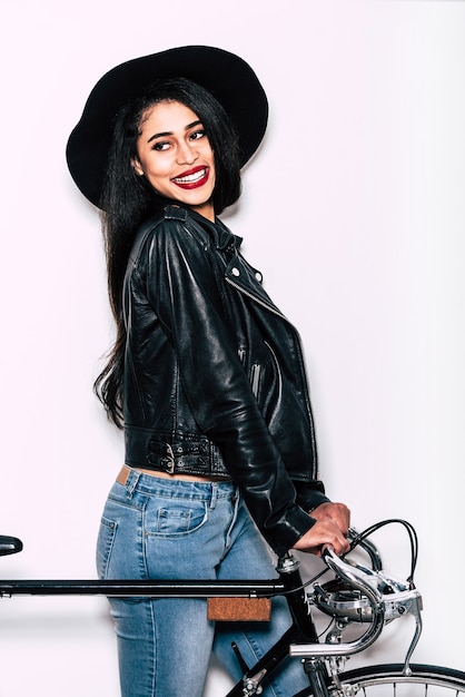 Ready to have some fun. Beautiful young mixed race woman in leather jacket and hat standing near black bicycle and looking away with smile