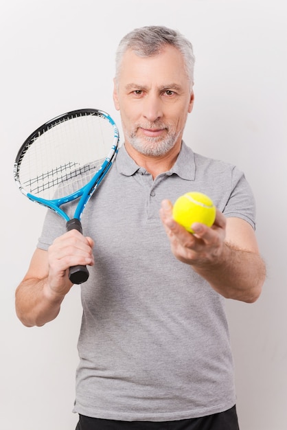 플레이할 준비가 되었나요? 흰색 배경에 서 있는 동안 테니스 라켓을 들고 테니스 공을 주는 자신감 있는 회색 머리 노인