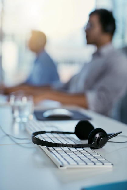 Foto pronto a fornire l'assistenza di cui hai bisogno primo piano di un auricolare che giace su una tastiera in un ufficio con gli agenti del call center in background