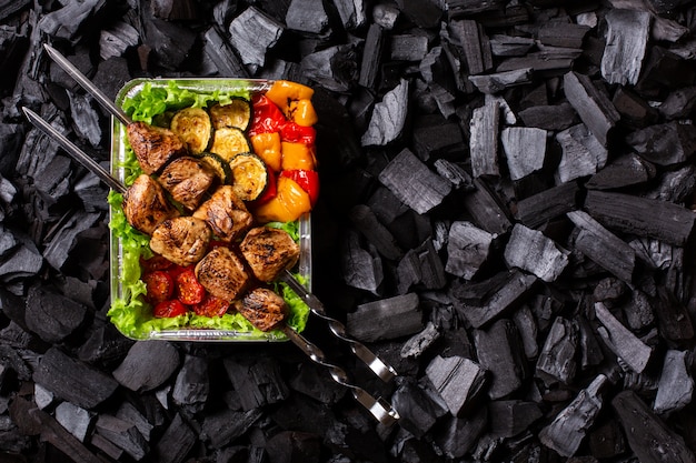 Ready shish kebab. Portion of grilled meat and vegetables in a disposable container on charcoal background. Copy space.