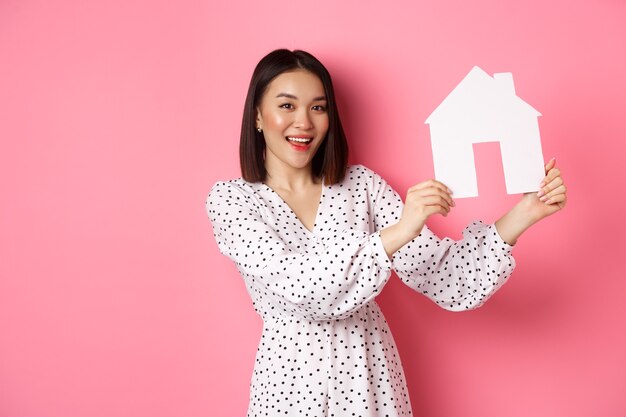 Real estate. Beautiful asian woman demonstrating paper house model, looking at camera confident, advertising home for sale, standing over pink