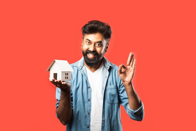 Real estate buying and people concept. Indian bearded man with miniature house model, keys and piggy bank on red