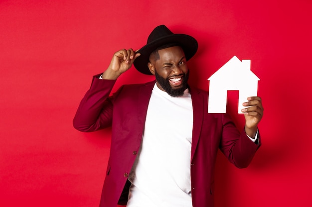 Недвижимость. Веселый темнокожий мужчина показывает бумажный домик и улыбается, рекомендуя брокера, стоя на красном фоне