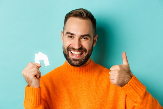 Real estate. Close-up of satisfied man showing thumb-up and small paper house maket, smiling pleased, standing over light blue background.