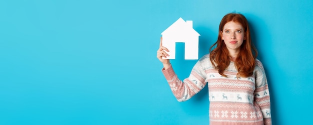 Photo real estate concept image of cute redhead girl looking curious at paper house model thinking of buyi