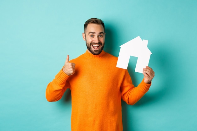 Real estate concept. Satisfied man recommending agency, showing thumb up and house maket, standing over blue background.