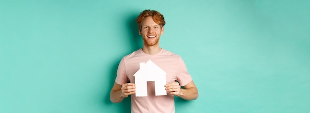 Real estate concept young man with red hair wearing tshirt showing paper house cutout and smiling ha