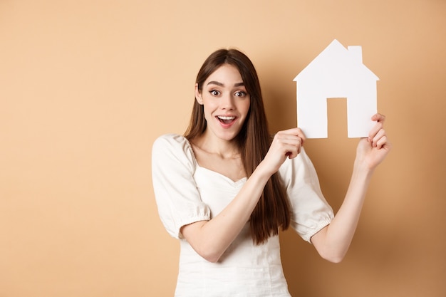 Immobiliare. la donna eccitata ha ottenuto un nuovo appartamento, mostrando il ritaglio della casa di carta e sorridendo felice, in piedi sul beige.