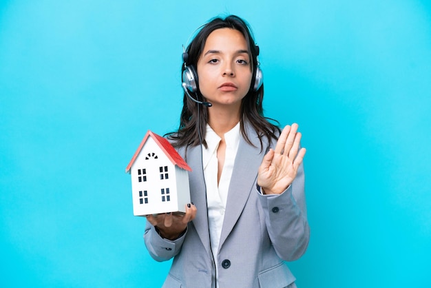 Real estate hispanic agent holding a toy house isolated on blue\
background making stop gesture