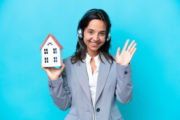 Real estate hispanic agent holding a toy house isolated on blue background saluting with hand with happy expression