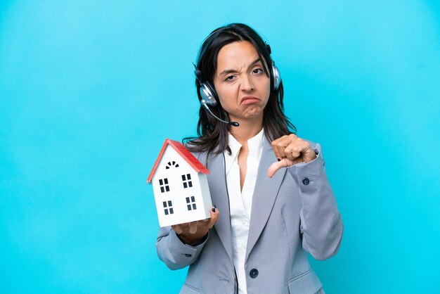 Real estate hispanic agent holding a toy house isolated on blue background showing thumb down with negative expression