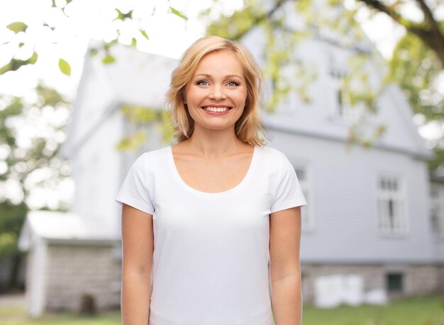 real estate, home and people concept - smiling woman in blank white t-shirt over private house background