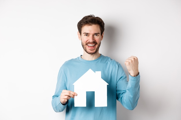 Real estate and insurance concept. Cheerful man buying property and celebrating, saying yes and showing paper house cutout, standing on white background.