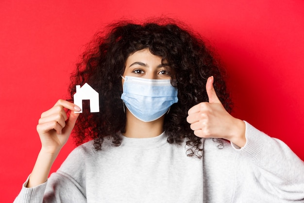 Real estate and pandemic concept. Close-up of woman recommending agency, wearing medical mask, showing thumbs up and paper house cutout, red background.