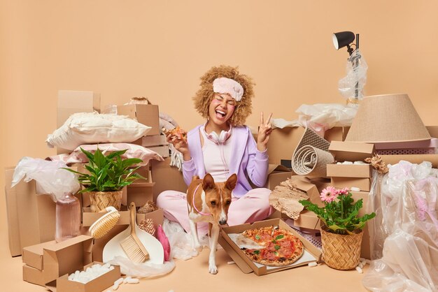Real estate purchase happy carefree european woman eats pizza
makes peace gesture poses around cardboard boxes with stuff on
floor near pet buys new property isolated over beige
background