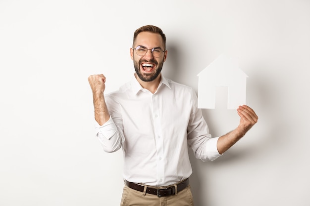 Real estate. Satisfied man rejoicing of founding perfect home apartment, holding paper house model, standing over white background.