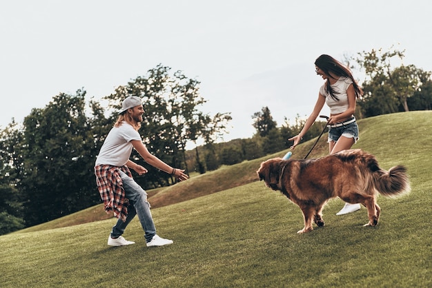 本当に楽しい。屋外で時間を過ごしながら犬と遊ぶ美しい若いカップルの全長