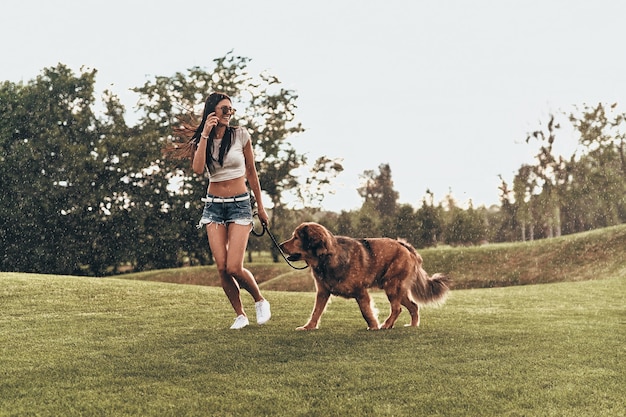 周りにいることの本当の喜び。彼女の犬と遊んで、屋外で実行しながら笑顔の美しい若い女性の完全な長さ