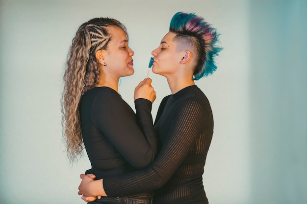 Real lesbian couple. Ethnic homosexual girlfriend playing with heart shaped lollipop to kiss a beautiful punk woman with blue mohawk hairdo.