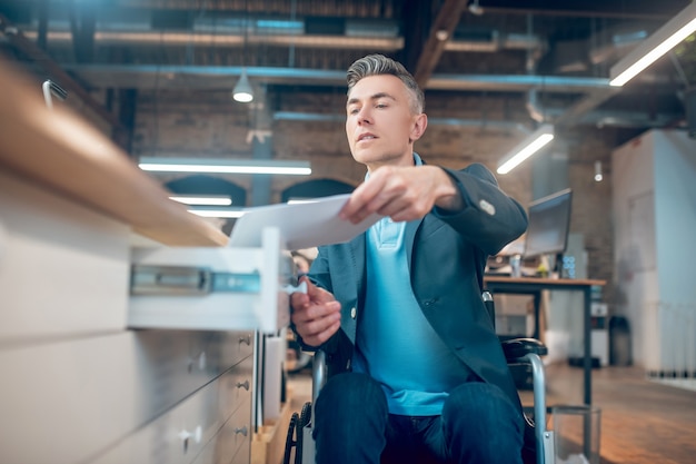 Real life. Young adult serious man in wheelchair with papers near open desk drawer in office space