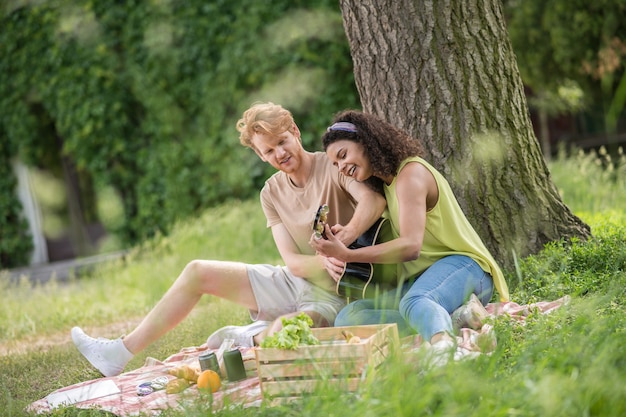 Real life. Young cheerful interested red-haired guy and mulatto girlfriend with guitar spending interesting leisure time on picnic in park