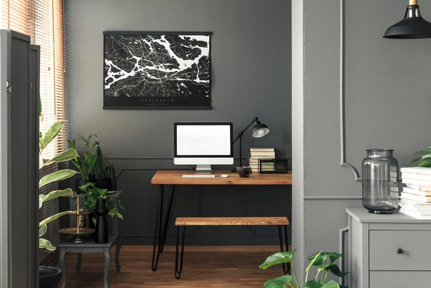 Real photo of dark open space living room interior with map poster on the wall above wooden desk for remote work with mockup computer books and lamp