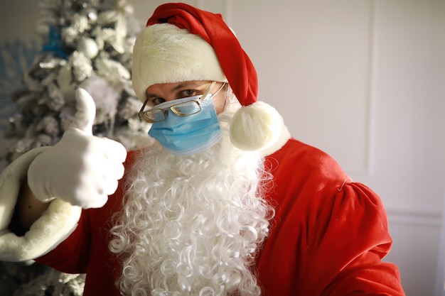 本物のサンタクロースと背景にクリスマスツリー、保護マスク、眼鏡、帽子を身に着けています。社会的距離のあるクリスマス。 COVID-19
