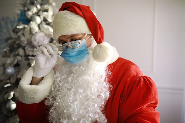 Real Santa Claus and christmas tree on a background, wearing a protective mask, glasses and hat. Christmas with social distance. Covid-19