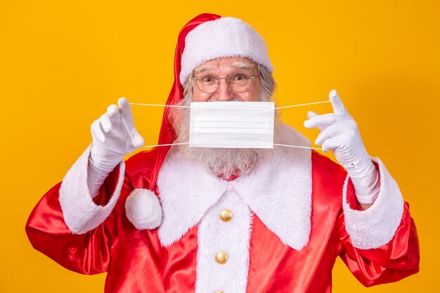 The real Santa Claus with a yellow background, holding a protective mask, glasses and hat. Christmas with social distance. Covid-19