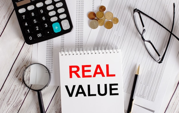 REAL VALUE written in a white notepad near a calculator, cash, glasses, a magnifying glass and a pen