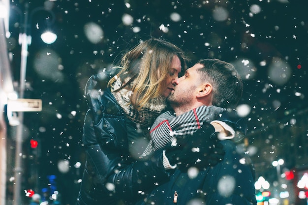 本当の若いカップルが雪の中で夜の街を一緒に歩いて、キス