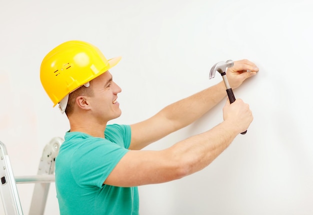концепция ремонта, строительства и ремонта дома - улыбающийся мужчина в желтом защитном шлеме забивает гвоздь в стену