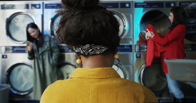 ランドリーサービスルームでアフリカ系アメリカ人のスタイリッシュな女性の後部。小さな洗濯場の混血の女性クライアント。座っていると洗濯機の作業と服のクリーニング中に待っている女の子の背面図