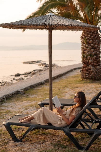 Rear angle view of lady in sunglasses resting on deck chair under straw beach parasol and reading novel Female tourist in summer attire enjoying holidays in Brac Island and spending time at seashore