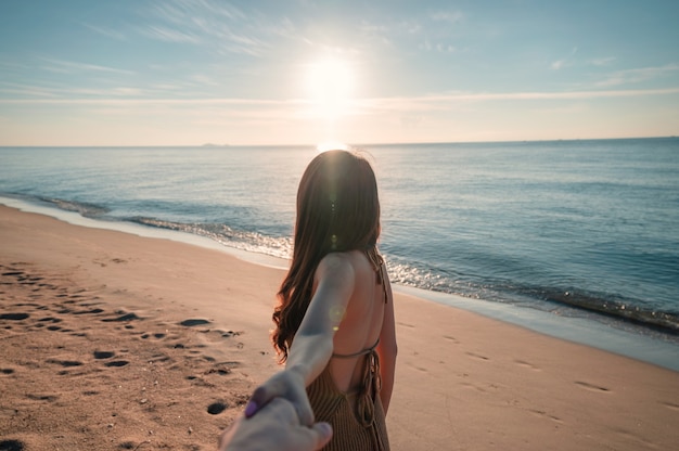 Parte posteriore della donna asiatica che si tiene per mano con il ragazzo che cammina sulla spiaggia con il sole al mattino durante le vacanze estive