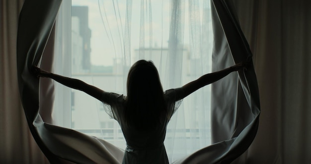 Rear back view young woman opening curtain lace standing in apartment home or modern hotel looking through window enjoying wellbeing light and city in the morning